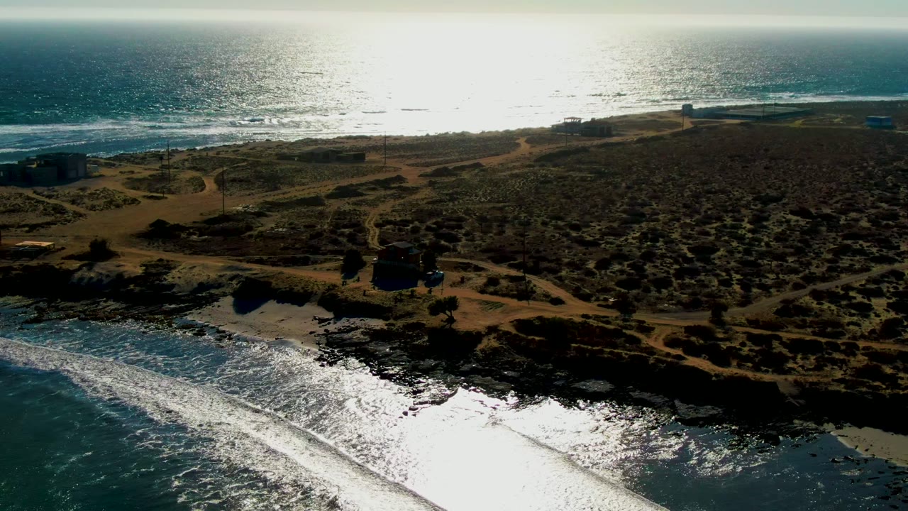 Aerial frame over La Bocana, Baja California Sur showing bright surf in the foreground, rocky shore, sandy tracks, scrub-covered dunes, distant small structures, and no visible people.
