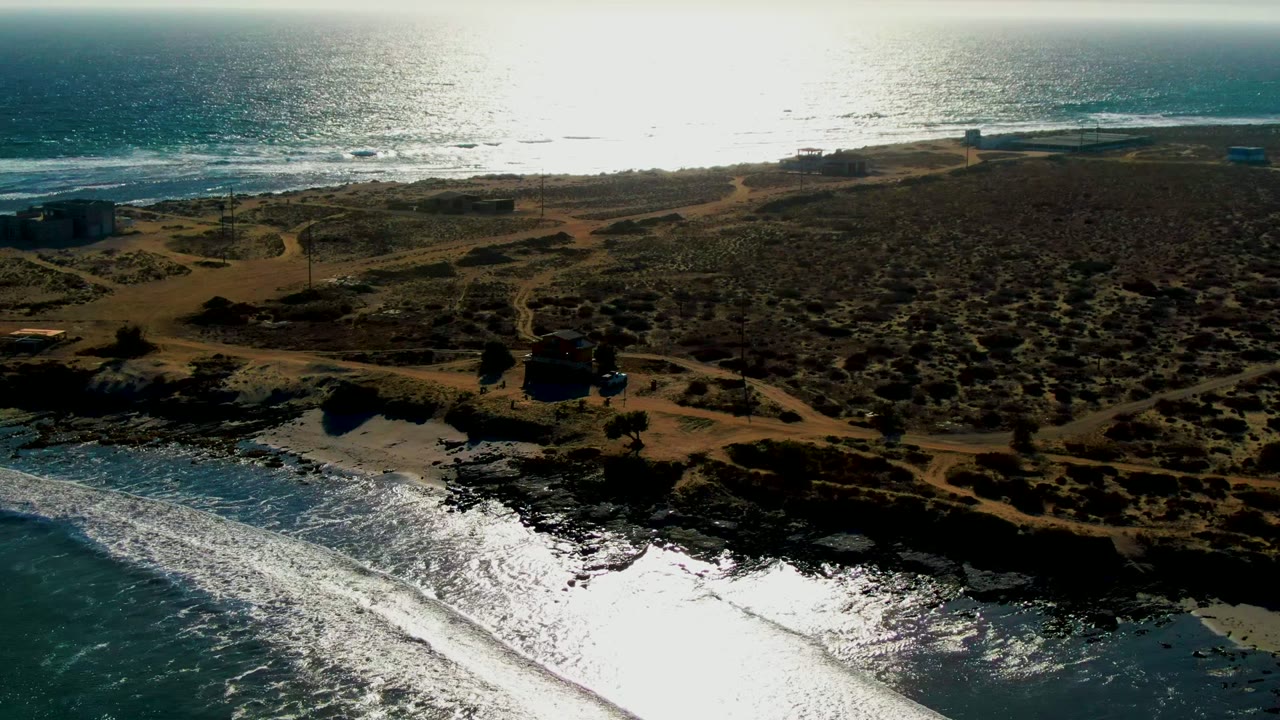 Aerial view in La Bocana with white wave bands along the lower edge, dark rocky coast, tan dirt tracks through low vegetation, small distant buildings, and no visible people.