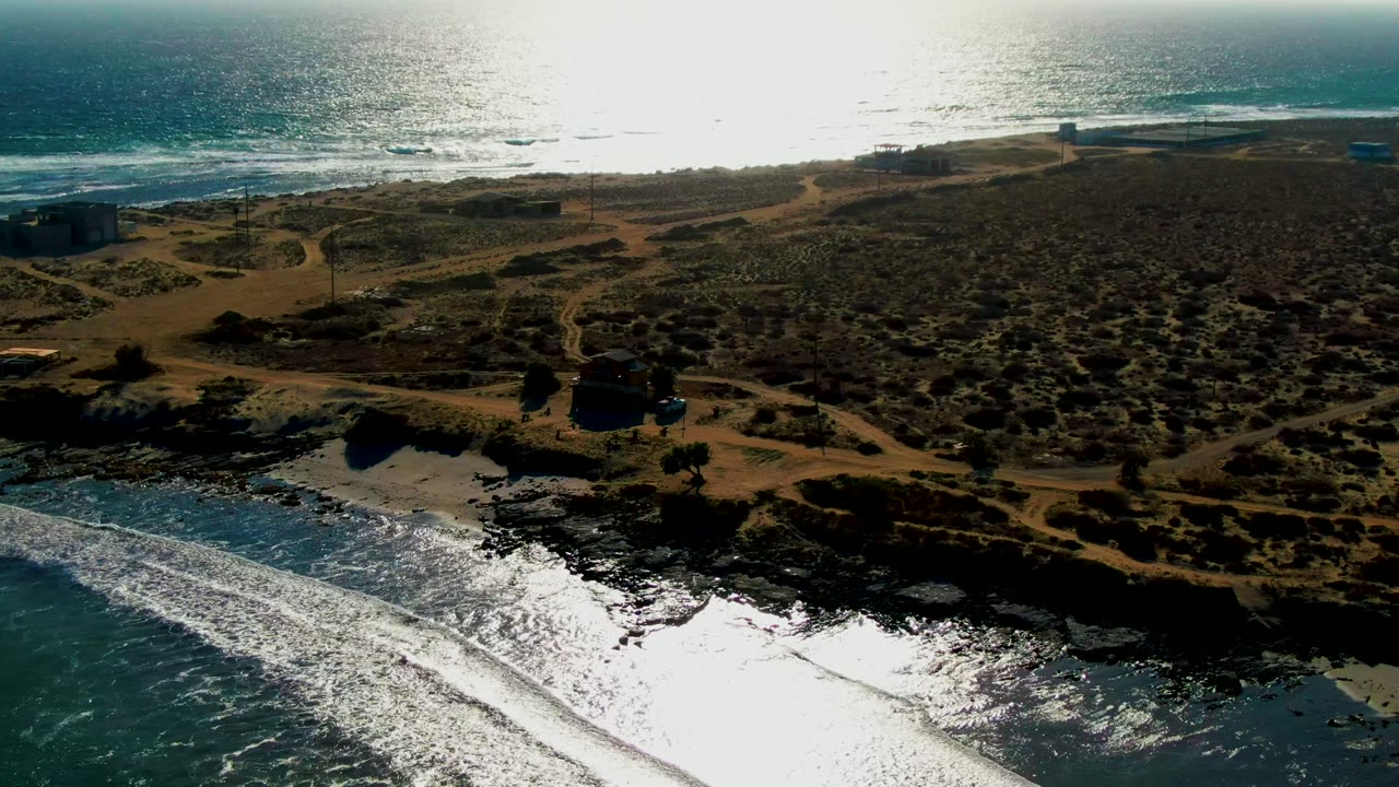 High aerial frame over La Bocana showing a reflective ocean horizon, foamy waves below, scrubby dunes crossed by sandy roads, distant coastal structures, and no visible people.