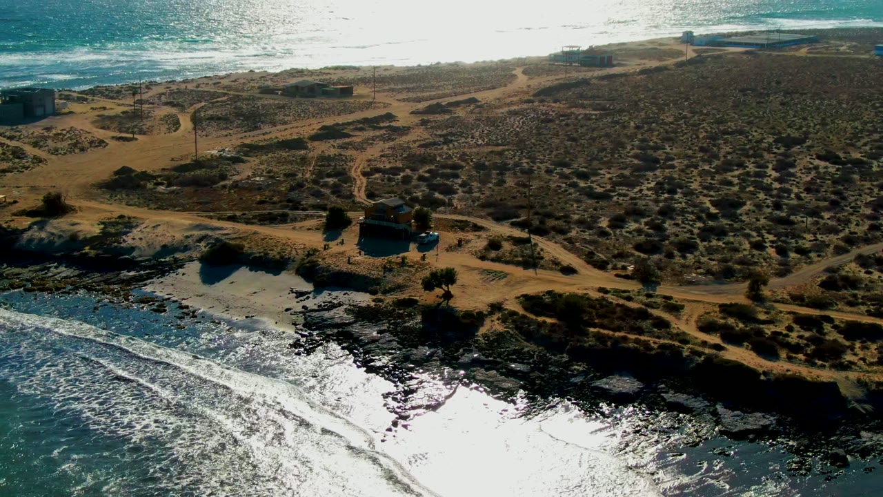 Aerial frame over La Bocana, Baja California Sur, with bright surf in the foreground, dark rocks along the shore, sandy dunes, dry vegetation, dirt tracks, and no visible people.