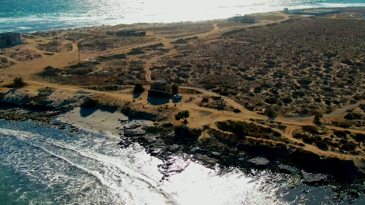 Drone view of waves, black rocks, dunes, and dirt paths near a small structure