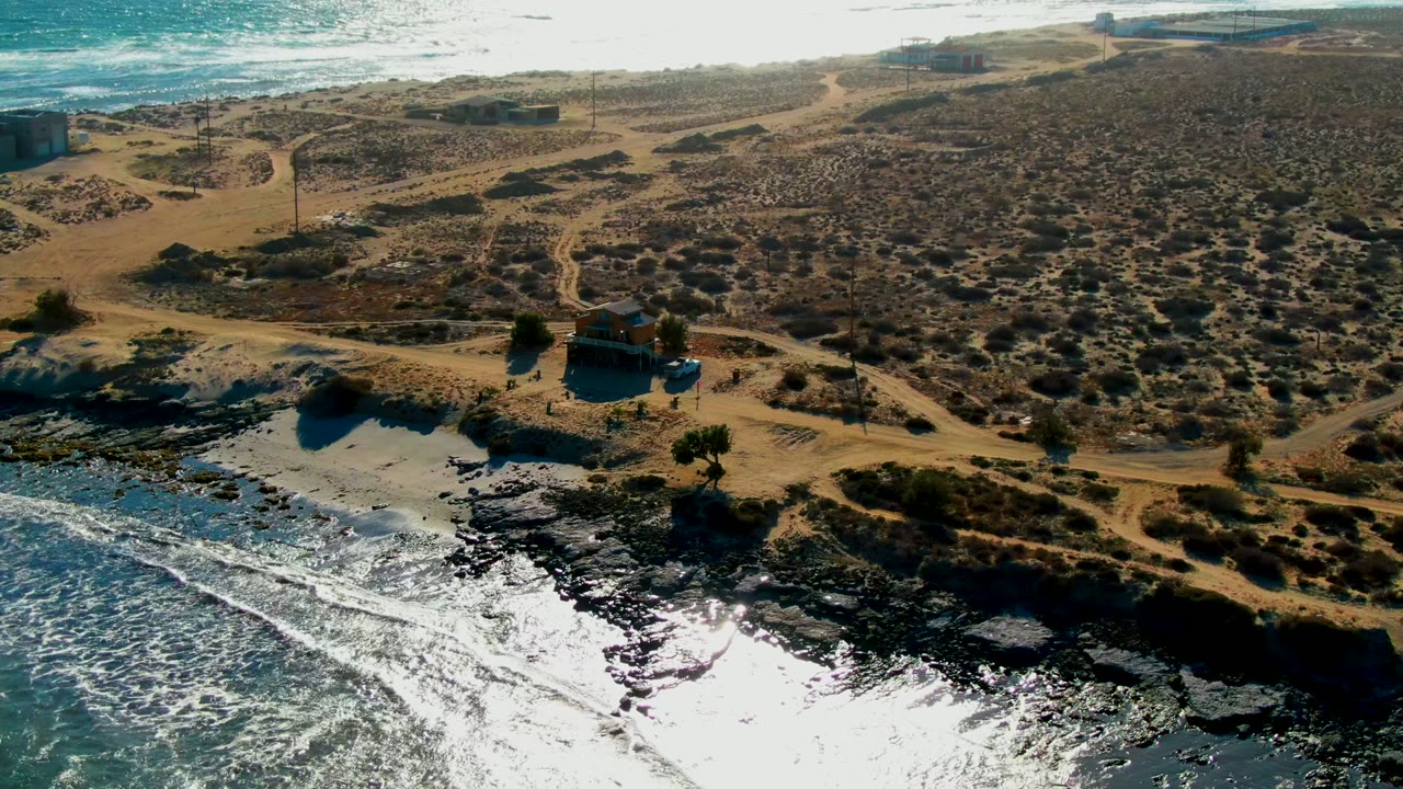 Drone-height frame over La Bocana with foamy ocean at the bottom left, a rocky beach edge, tan dunes crossed by dirt tracks, scattered scrub, utility poles, and no visible people.