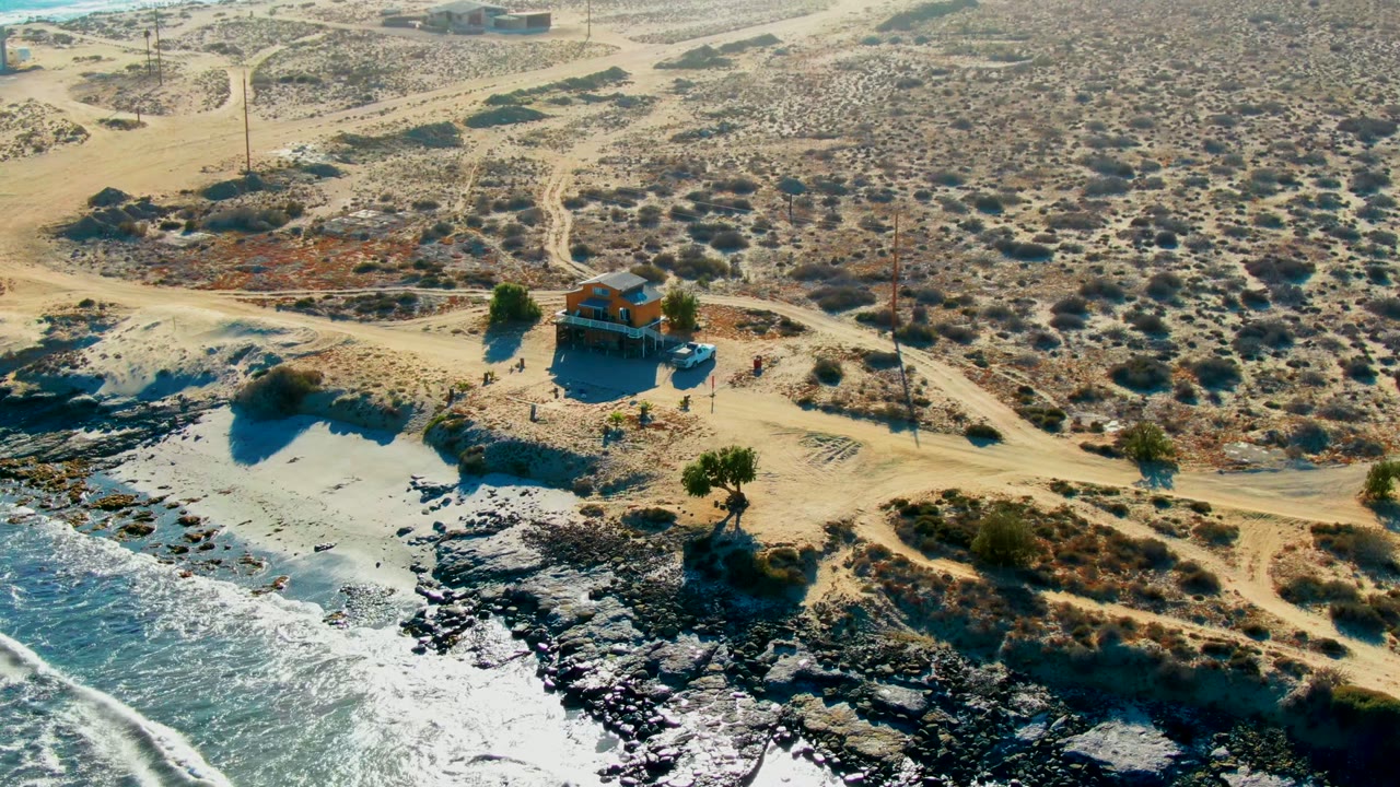 Aerial frame from La Bocana, Baja California Sur showing a small house beside dirt roads, sparse desert scrub, dark shoreline rocks, and ocean foam at the lower left.