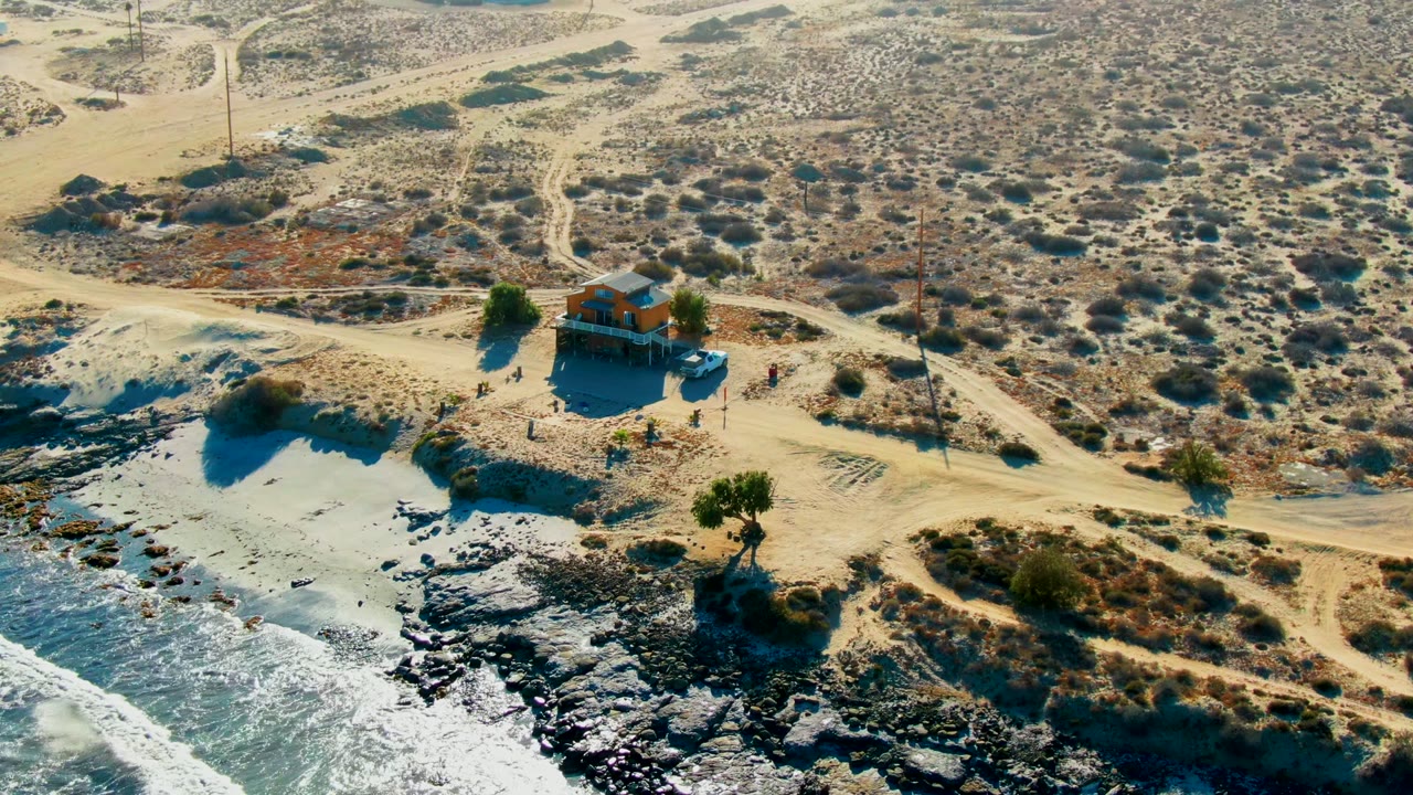 Aerial La Bocana frame showing the house slightly right of center, pale dirt tracks crossing sandy scrubland, dark coastal rocks, and white water breaking at the shore with no visible people.