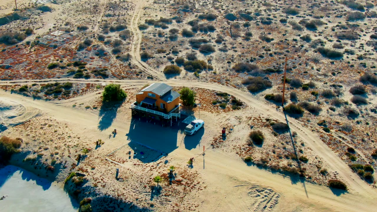 Opening aerial frame in La Bocana, Baja California Sur showing a small orange desert building, a white vehicle, dirt roads, scrub vegetation, utility poles, long shadows, and visible people nearby.