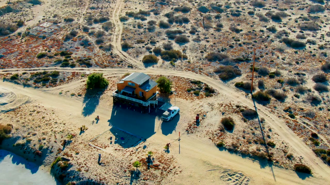 High aerial view of a small building, parked vehicle, people, dirt roads, and desert scrub.
