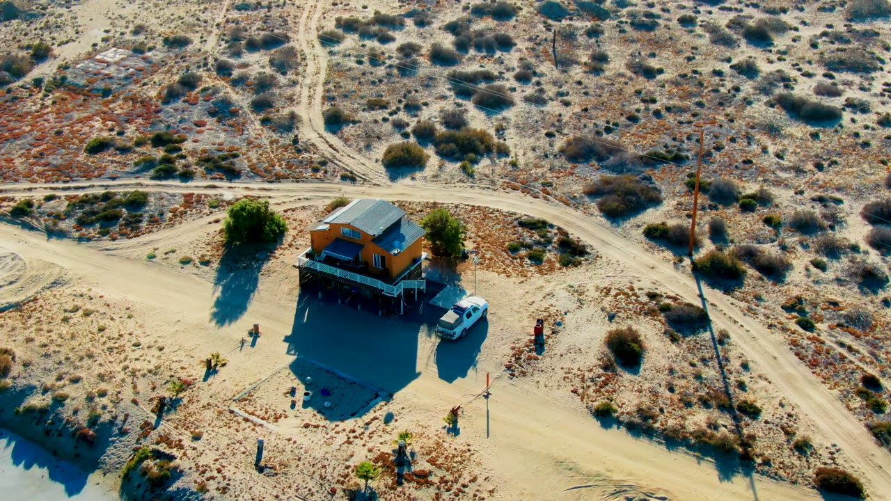 At 0:02, the La Bocana frame shows the small building casting a broad shadow, a white vehicle parked beside it, several visible people, crossing dirt roads, and low desert vegetation.