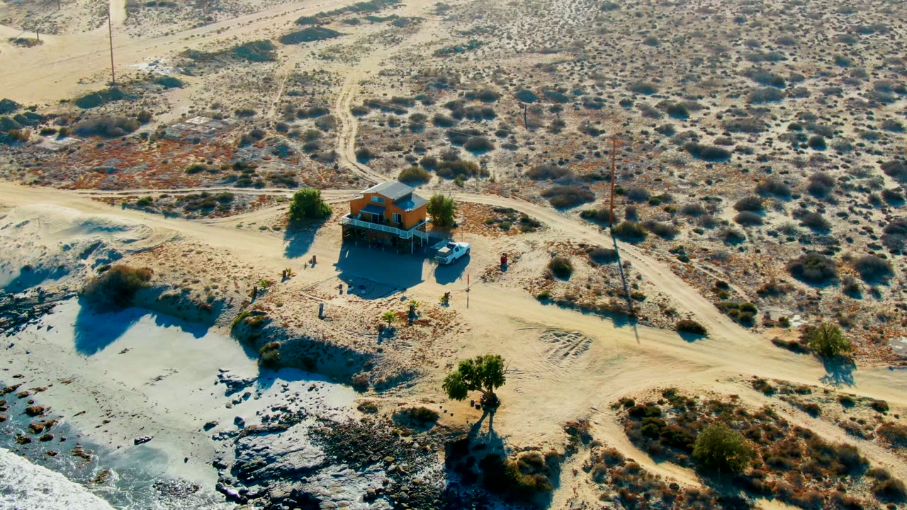 Aerial frame from La Bocana, Baja California Sur showing a small raised building, pale dirt roads, scrubby desert ground, a blue vehicle, and tiny visible people near the structure.