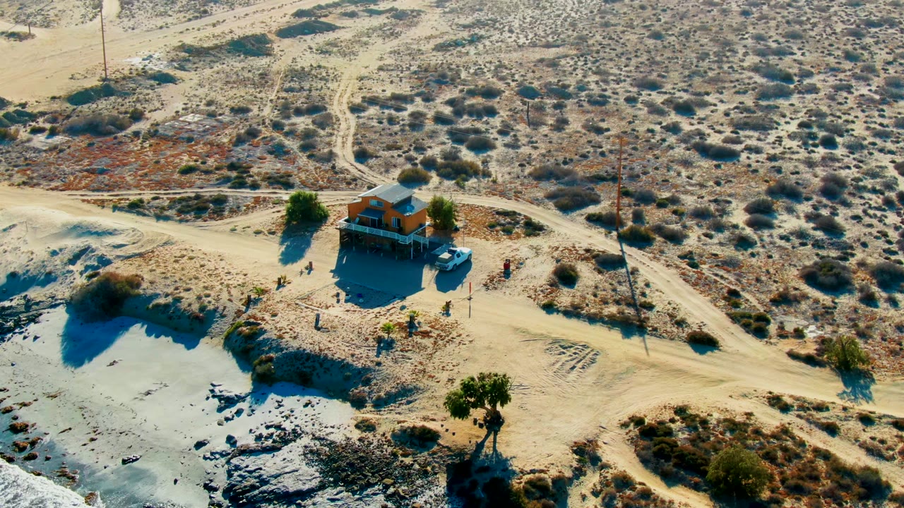 High aerial view in La Bocana with the small building slightly lower in frame, the blue vehicle parked beside it, people as tiny figures, and dirt tracks crossing the scrubland behind.