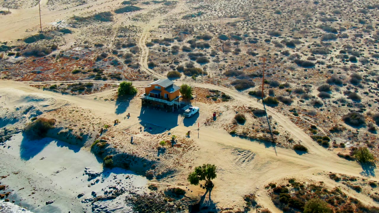 Aerial La Bocana frame showing the shack near the lower center, a road bending across the scene, scrub-covered desert behind it, and tiny people visible around the building area.