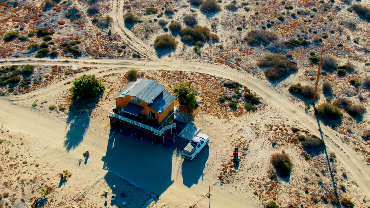 Overhead frame in La Bocana, Baja California Sur showing a small orange desert house, a white car beside it, several people around the lot, and pale dirt roads through dry scrub.