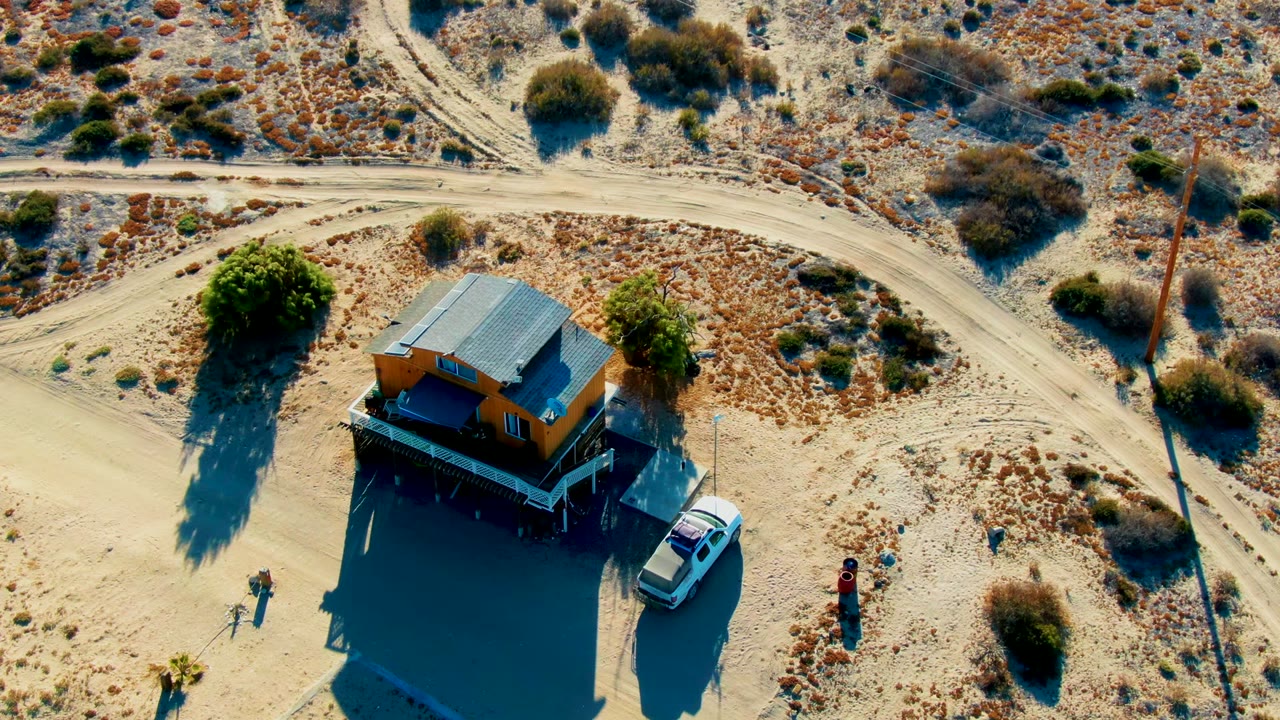 Aerial frame in La Bocana, Baja California Sur showing the orange house shifted to the right of center, the white car along its side, people nearby, and tire-marked sand around the road.