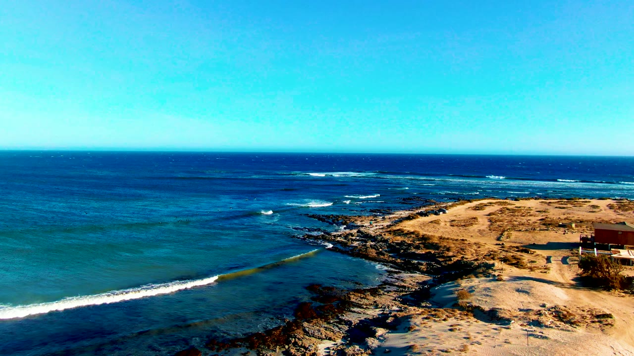 Wide aerial view over La Bocana, Baja California Sur, with blue ocean, white surf, rocky shoreline, pale sand, clear sky, and no visible people.