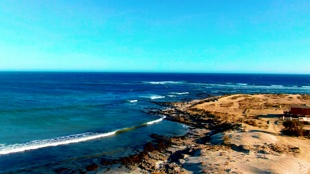 Aerial coastal view with white surf curving toward dark rocks and sand.