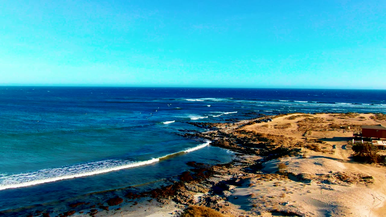 La Bocana aerial shoreline view with whitecaps spreading across blue water, a sandy point at right, dark rocks below, and no visible people.