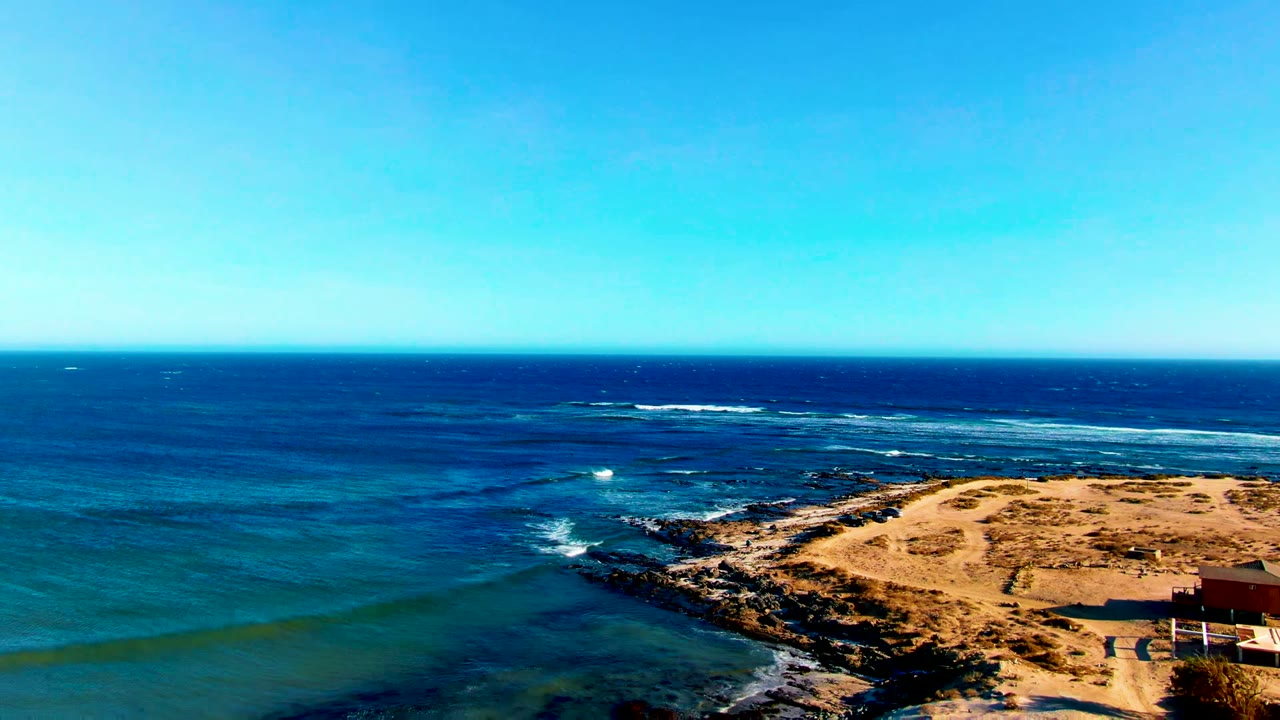 Opening frame over La Bocana, Baja California Sur, with blue ocean in the foreground, a rocky sandy point on the right, white surf offshore, and no visible people.