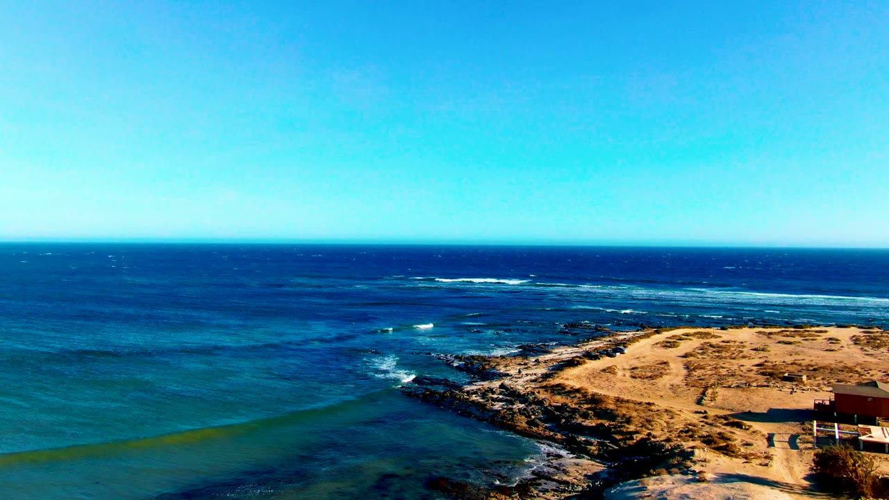 Blue water and white surf lines around a rocky point beside sand.