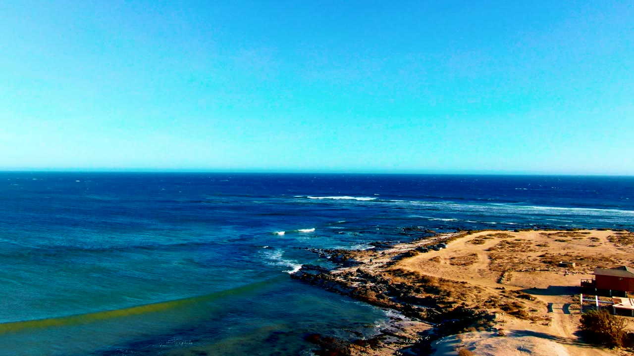 At 0:02 in La Bocana, turquoise nearshore water and darker offshore water surround the rocky sandy point, with a straight horizon and no visible people.