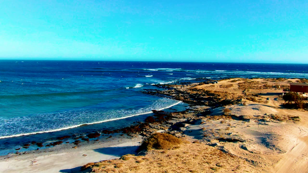 Elevated view over La Bocana, Baja California Sur with turquoise water, low waves, sandy beach, rocky outcrops, dry coastal ground, and no visible people.