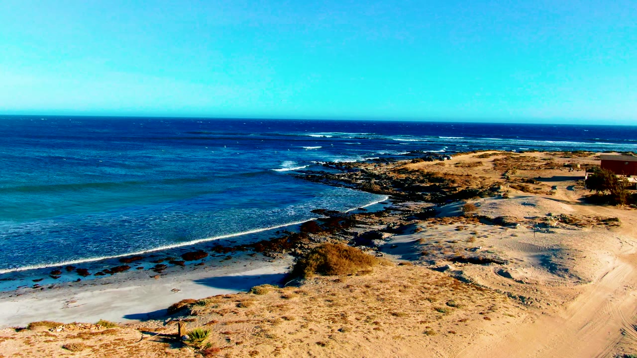 Wide elevated frame of La Bocana showing bright blue water, a pale sandy beach, dark rocky shoreline sections, dry tan ground, and no visible people.