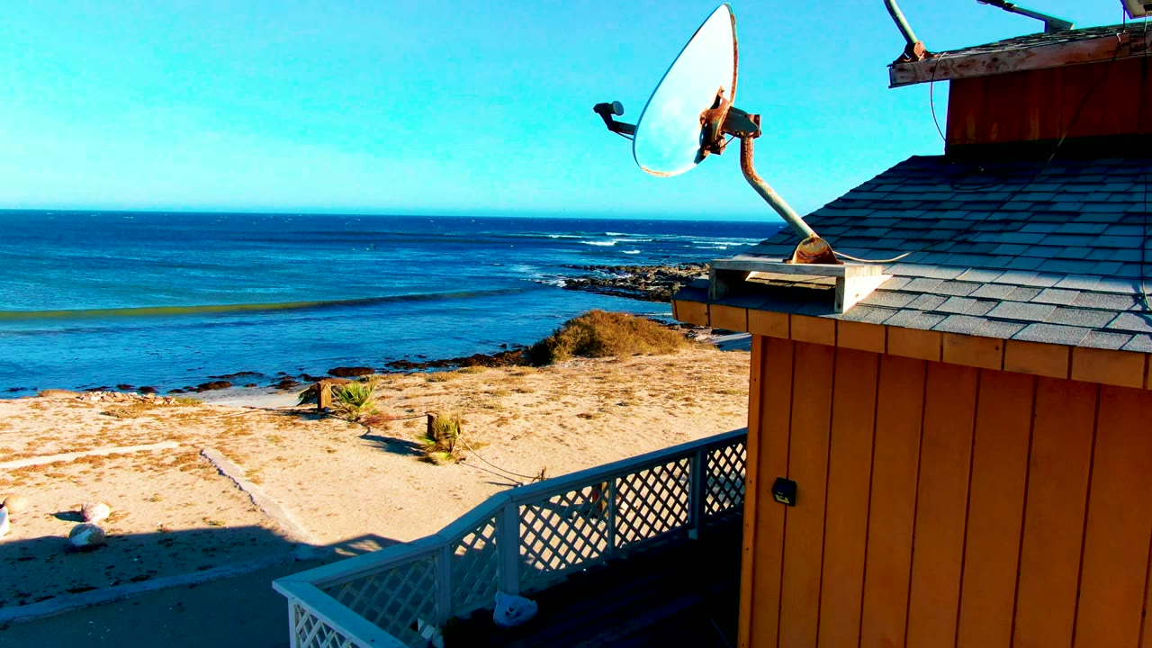 At 0:00 in La Bocana, Baja California Sur, a satellite dish and orange beach house roof edge overlook blue ocean, sand, and a white deck railing, with no visible people.