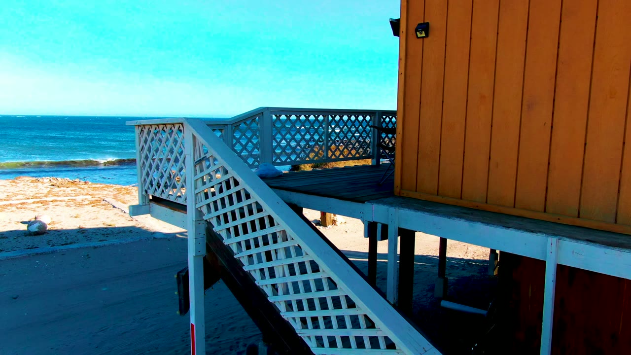 At 0:02 in La Bocana, Baja California Sur, white lattice stairs and deck railings fill the right side beside orange siding, while sand and blue ocean remain visible behind them, with no visible people.
