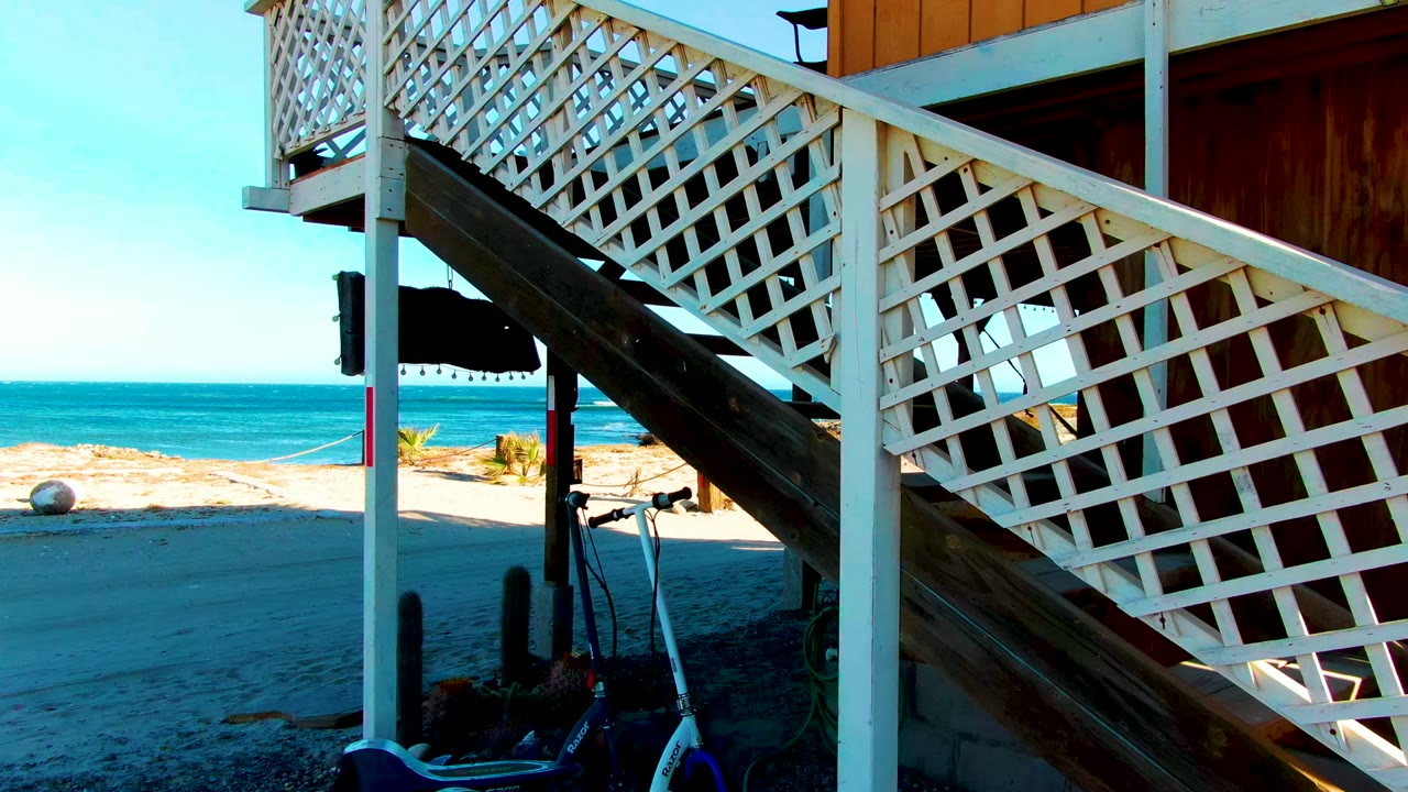 White lattice beach staircase in La Bocana, Baja California Sur, Mexico, with ocean and sand in the background, a scooter near the lower posts, and no visible people.