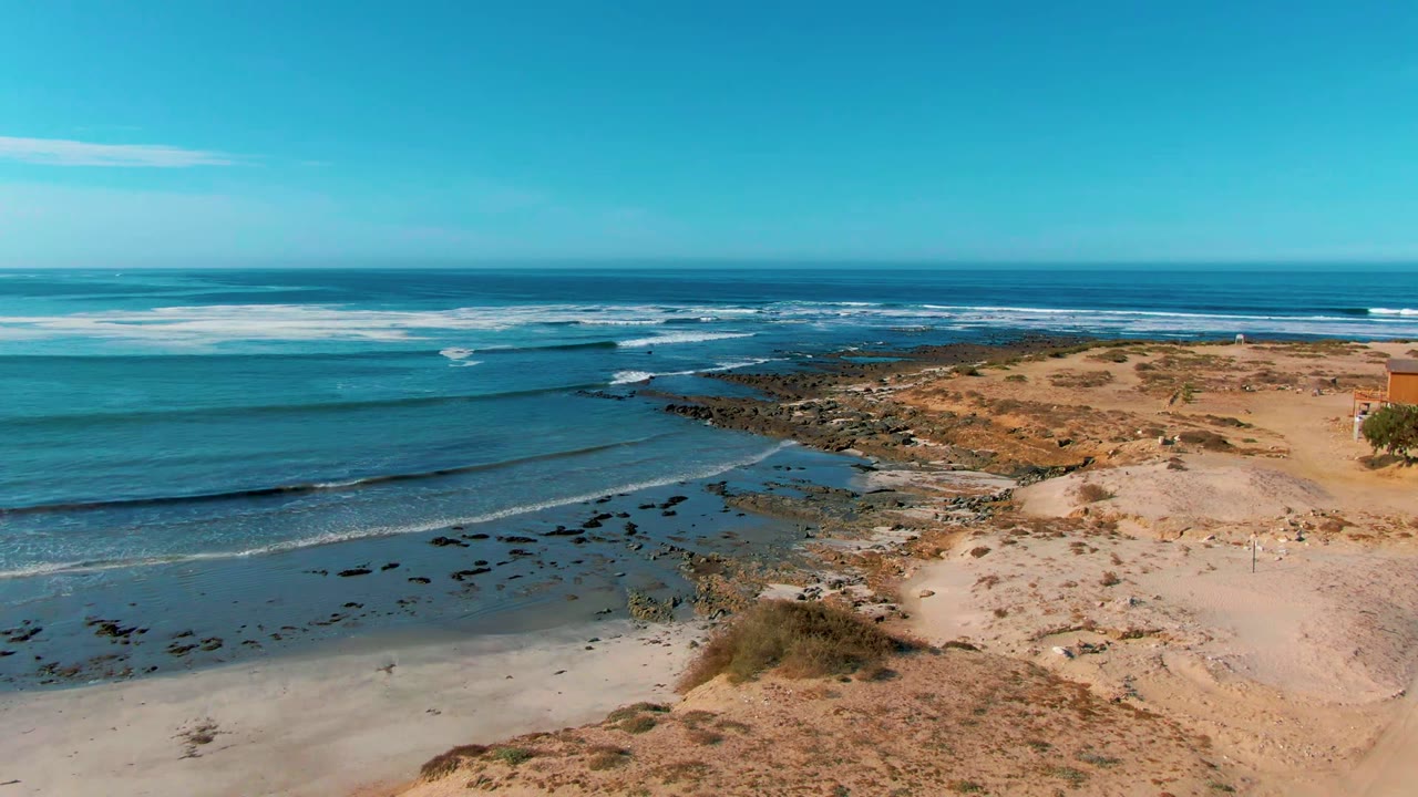 Aerial view over La Bocana, Baja California Sur, with blue water entering from the left, white surf lines near the horizon, sand and dark rocks along the shore, and no visible people.
