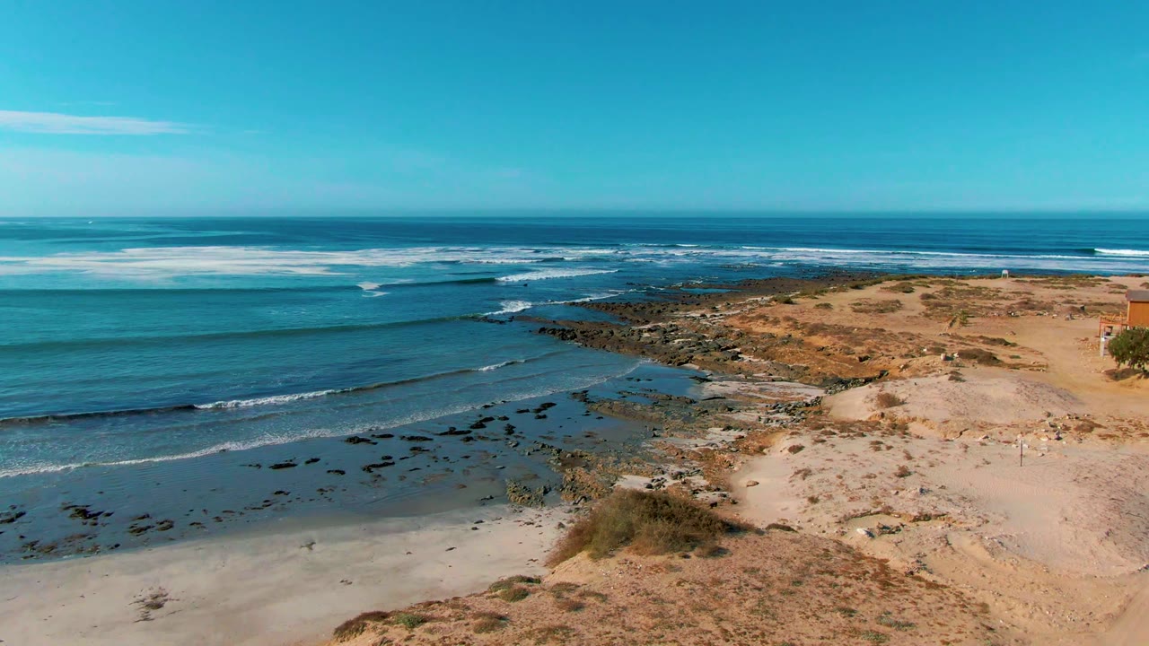 Waves roll toward shore beside dry dunes and dark rocks.