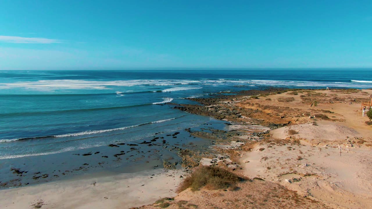 A wide aerial frame over La Bocana shows white foam bands across the blue ocean, a rocky shoreline in the middle distance, pale sand to the right, and no visible people.
