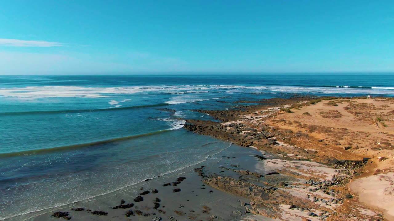 Aerial view of La Bocana, Baja California Sur, with blue ocean water moving toward dark rocks, tan sand, low dunes, a clear horizon, and no visible people.