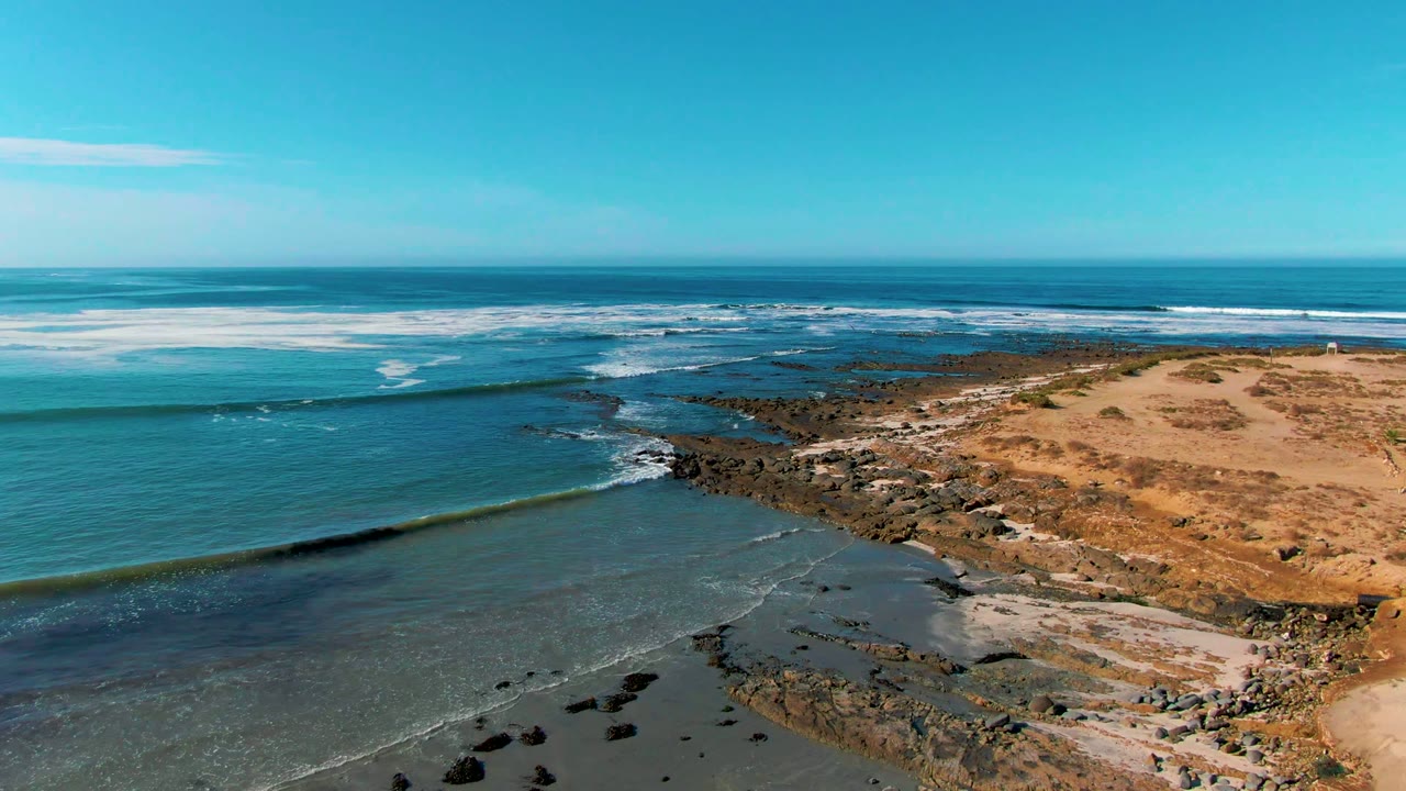 Ocean swell nearing dark rocks beside tan coastal dunes