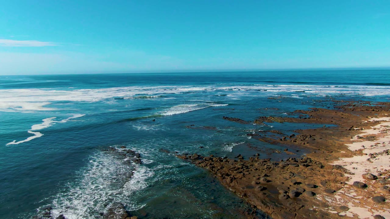 Aerial view in La Bocana, Baja California Sur, with turquoise water, white surf, dark rocks, sandy shoreline patches, and no visible people at the start of the clip.