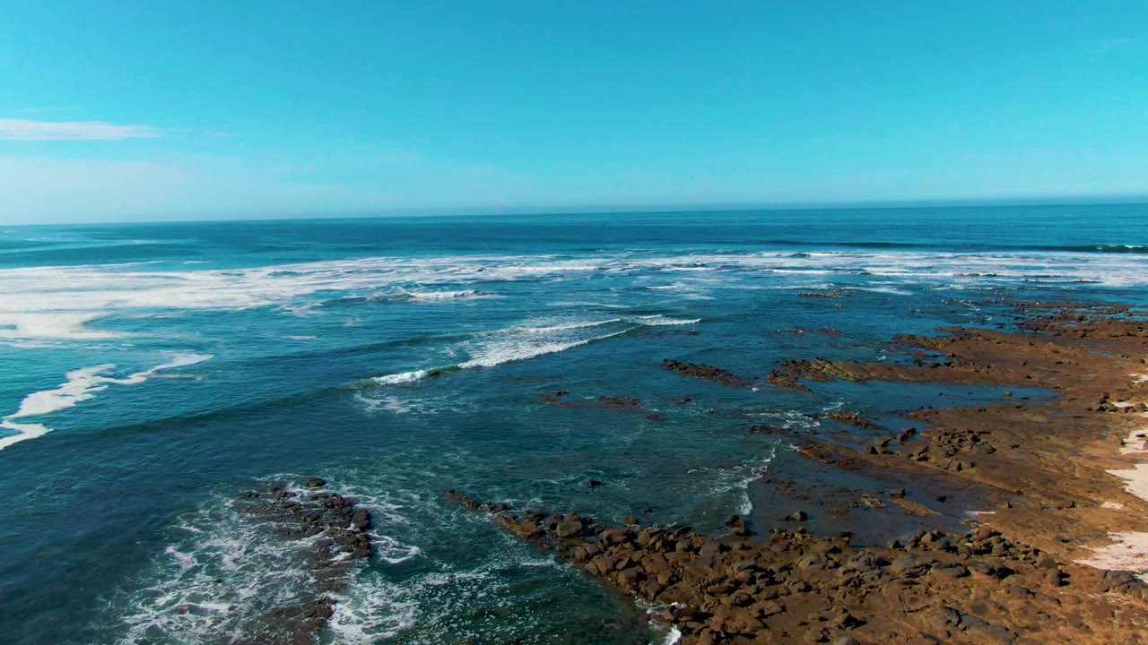 Aerial view in La Bocana, Baja California Sur, with white foam spreading across shallow reef water, rocky outcrops at lower frame, sandy right edge, and no visible people.