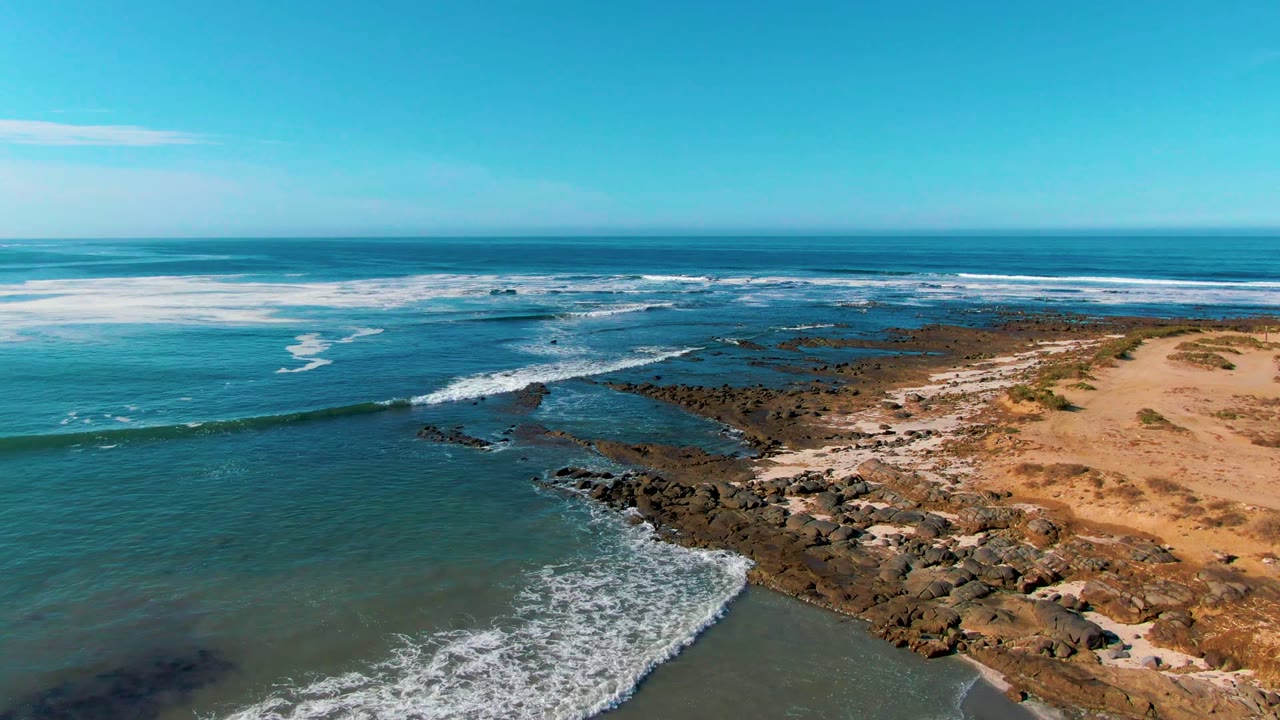 Aerial frame from La Bocana, Baja California Sur showing blue ocean, white surf curling over dark shoreline rocks, tan sand to the right, and no visible people.
