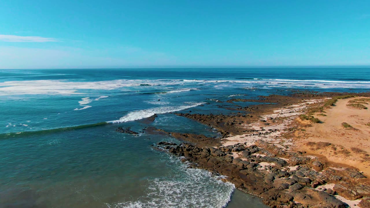 Blue ocean surf rolls toward a rocky point beside tan sand.