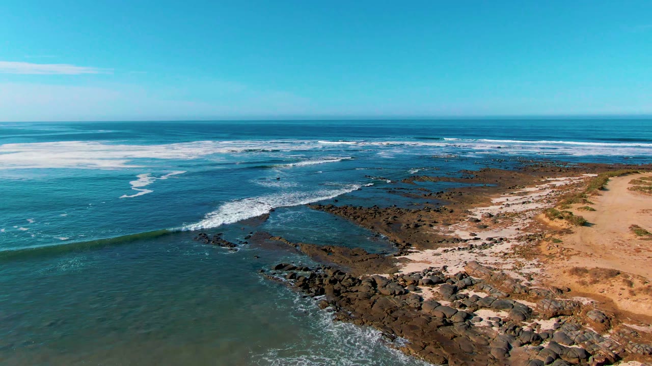 La Bocana aerial frame showing white foam spread over shallow dark rocks, open blue water beyond, tan sand and sparse coastal scrub to the right, and no visible people.