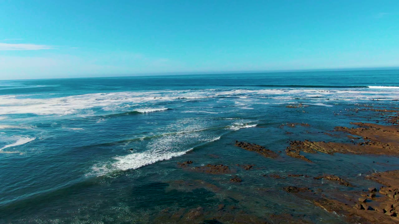 Aerial view at La Bocana, Baja California Sur, with white surf spreading over shallow rocky shoreline, open ocean on the horizon, and no visible people.