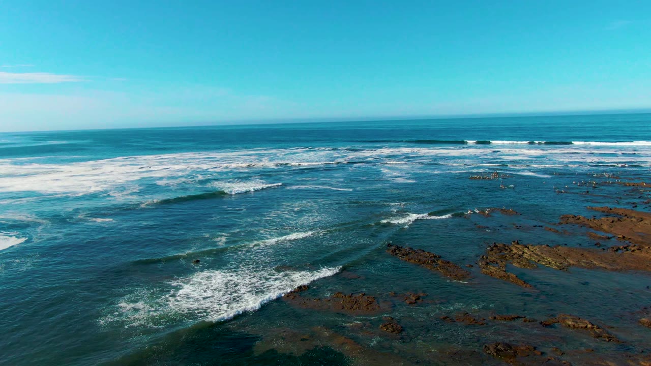 Aerial frame from La Bocana showing broken foam over dark coastal water, rock shelves clustered to the right, and a clear horizon with no visible people.