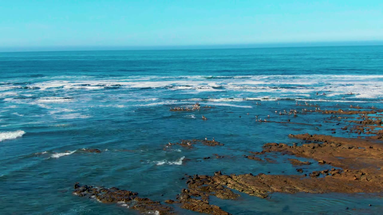 Wide elevated view of La Bocana ocean surf breaking in white bands beyond exposed brown rocks, with blue water and no visible people.