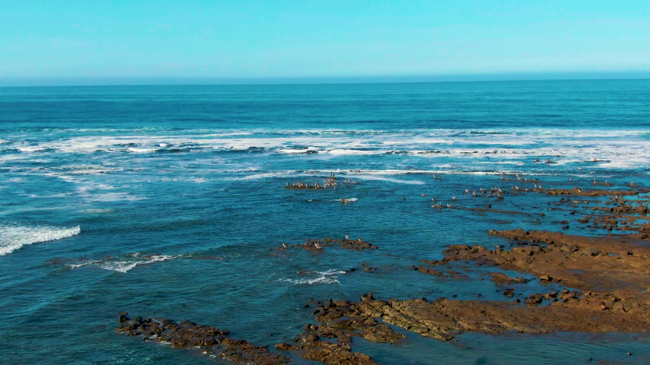 Elevated La Bocana shoreline frame with a rocky reef shelf on the right, scattered dark rocks in shallow water, and white breakers across the background, no visible people.