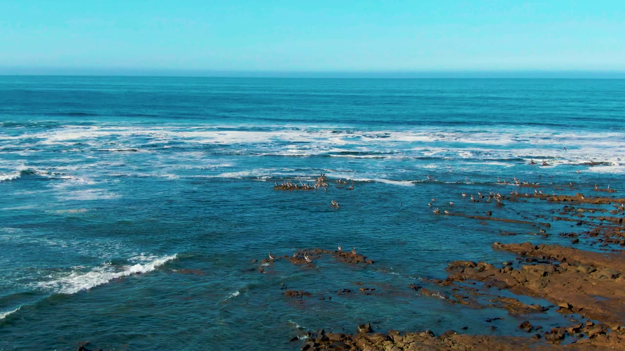 Wide elevated view of La Bocana ocean surf breaking across shallow rocky water, with brown rocks at the lower right and no visible people.