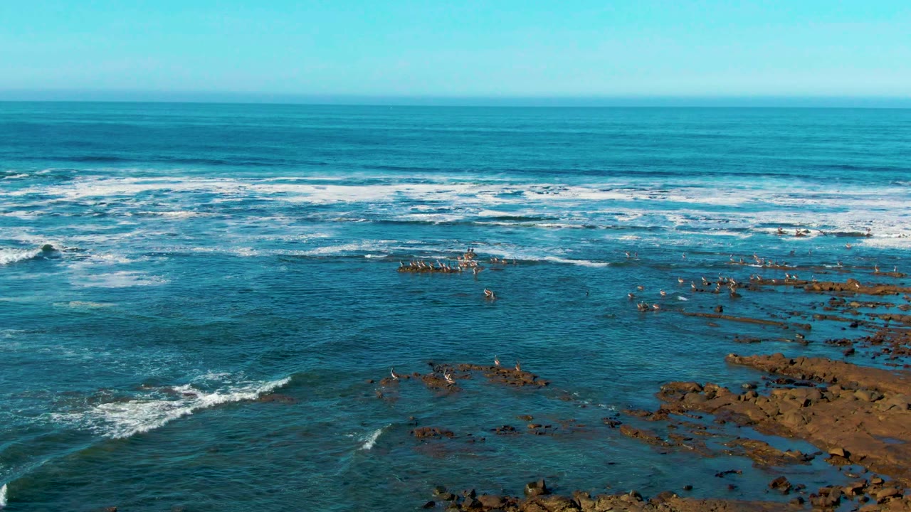 White surf rolls across blue water beside exposed coastal rocks.