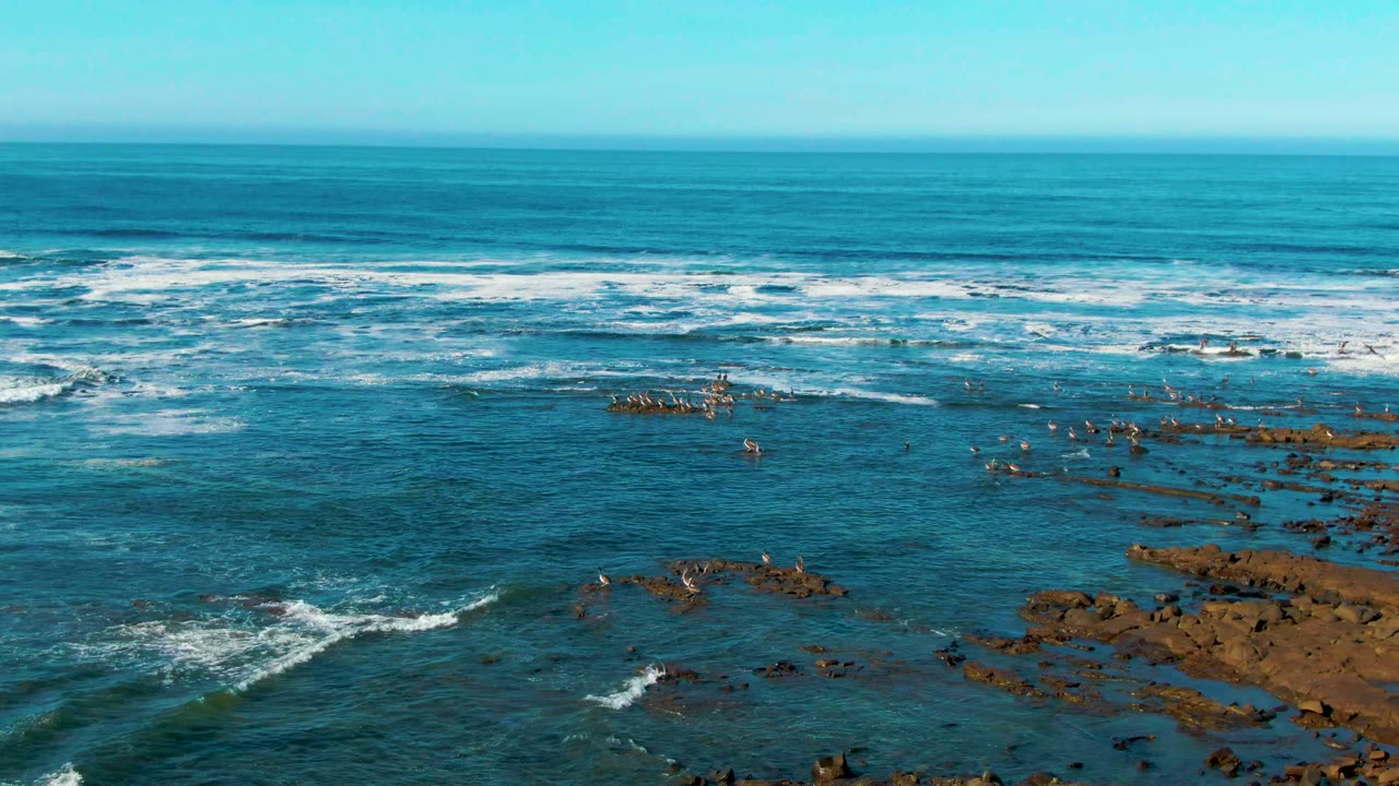 Elevated La Bocana coastal frame with layered wave breaks across the midground, darker rock patches in the shallows, and no visible people.