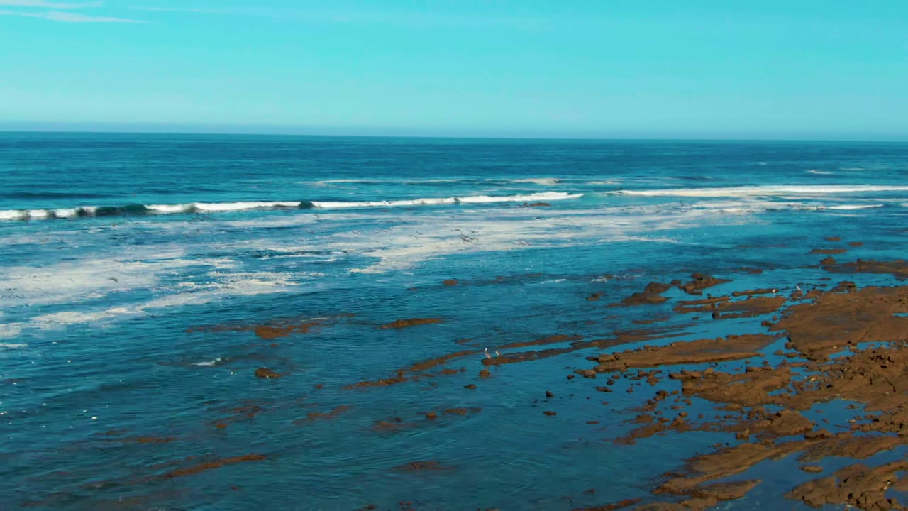 Aerial view in La Bocana, Baja California Sur, with blue ocean, white surf bands, brown rock flats at lower right, and no visible people.