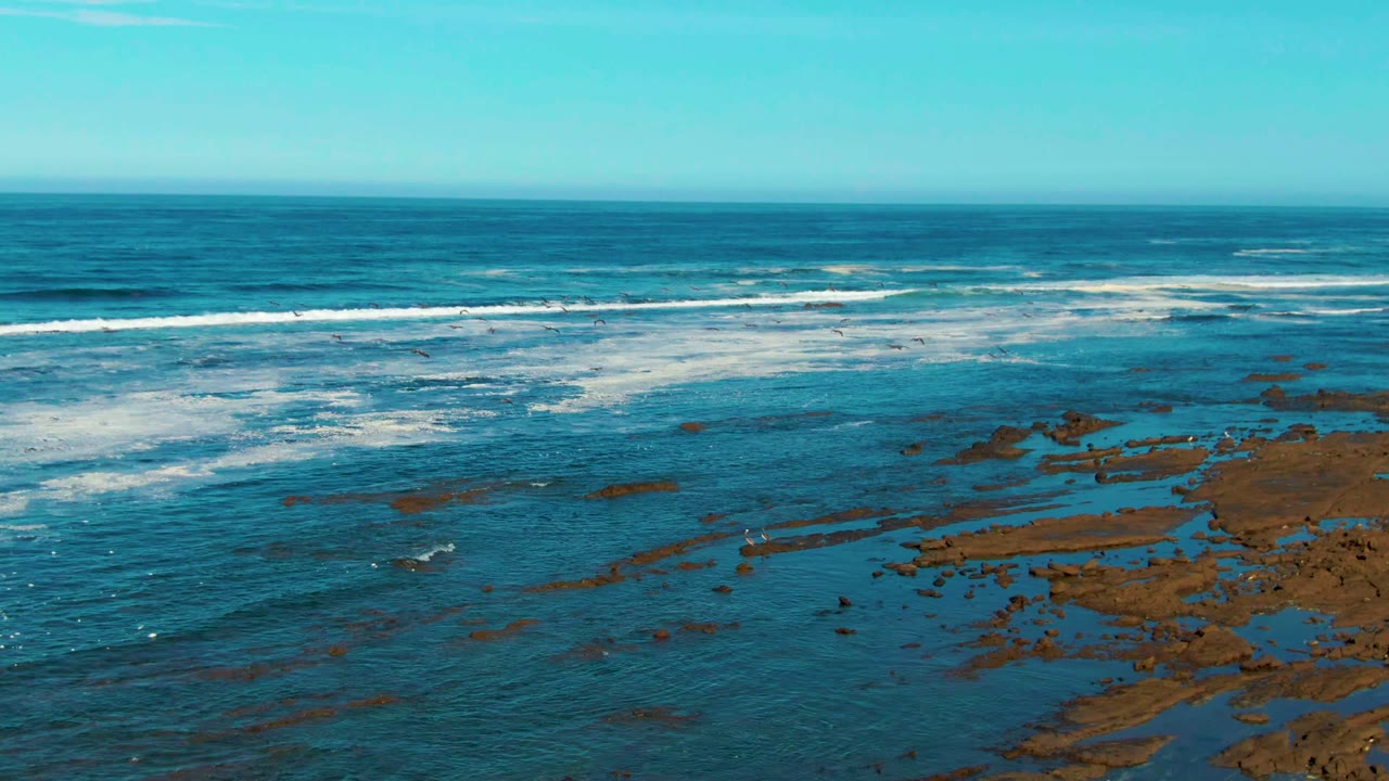 Aerial coastal view in La Bocana with white breakers crossing the midground, darker blue water offshore, rock patches at right, and no visible people.