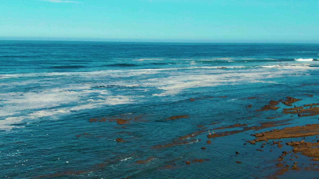 Aerial-looking view of blue ocean and white surf over rocky shallows at La Bocana, Baja California Sur, with a clear sky and no visible people.