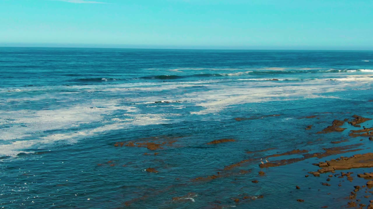 Rocky reef patches sit beneath shallow blue water while white breakers roll across the midground at La Bocana, Baja California Sur, with no visible people.