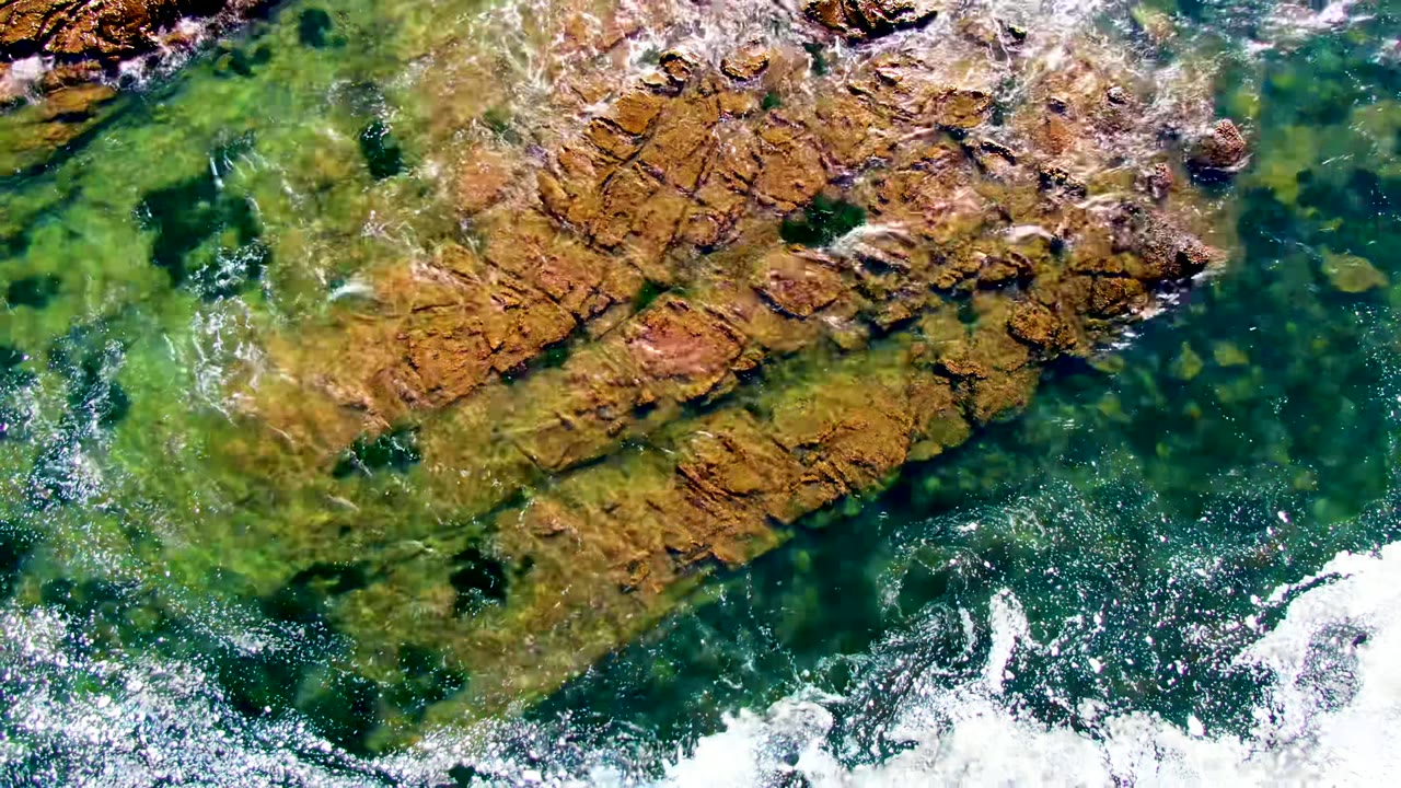 Overhead aerial frame at La Bocana showing clear green water crossing brown coastal rock shelves, bright white foam at the lower edge, and no visible people.