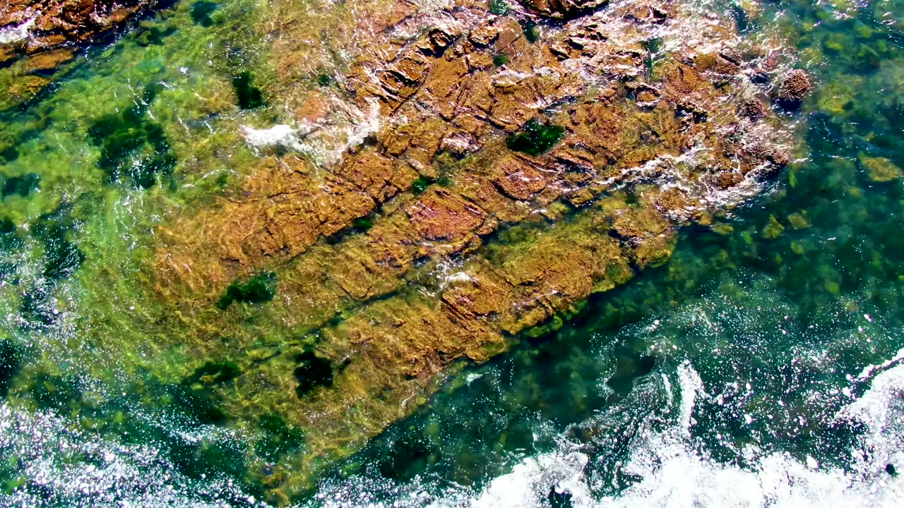 Top-down La Bocana shoreline frame with orange-brown rock near the upper half, clear teal water below, scattered white foam, and no visible people.